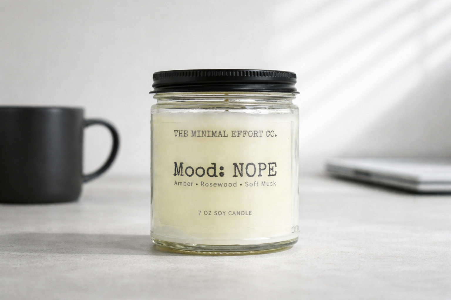 Candle labeled 'Mood: NOPE' by The Minimal Effort Co. on a table with a mug and phone.