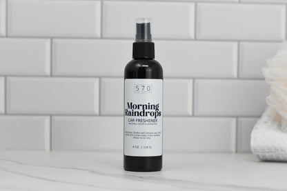 A spray bottle of Linen & Room Spray by 570 Soap Co., a natural odor eliminator, rests on a marble surface. The label is white with black text against a backdrop of white subway tiles, offering refreshing fragrances for your drive.