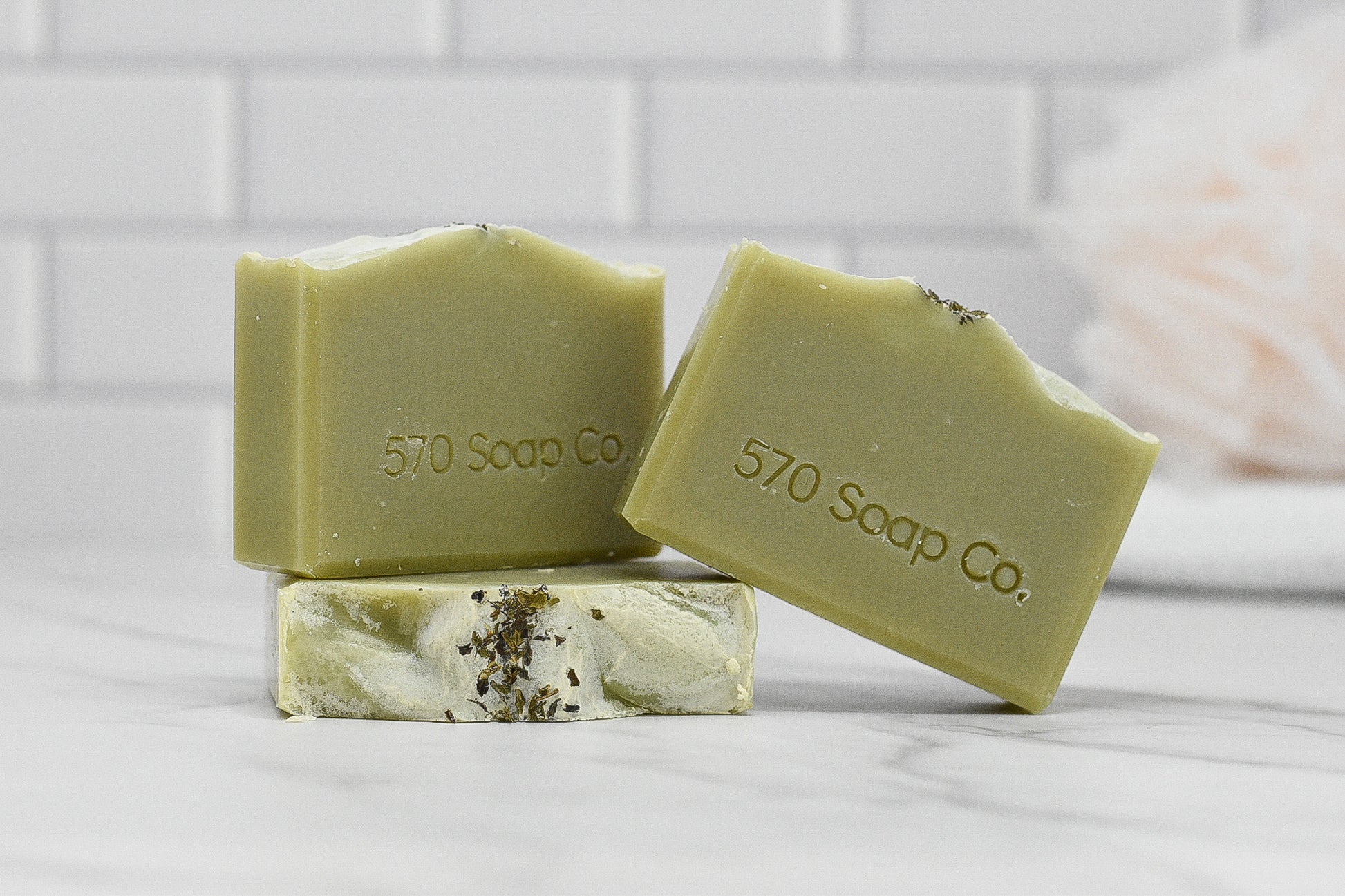 Three bars of the Aloe & Mint Bar Soap - All Natural from 570 Soap Co. are displayed on a marble surface. With their natural, earthy tones and dried herb toppings, these soaps invigorate the senses. A white, tiled wall serves as the backdrop.