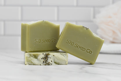 Three bars of the Aloe & Mint Bar Soap - All Natural from 570 Soap Co. are displayed on a marble surface. With their natural, earthy tones and dried herb toppings, these soaps invigorate the senses. A white, tiled wall serves as the backdrop.