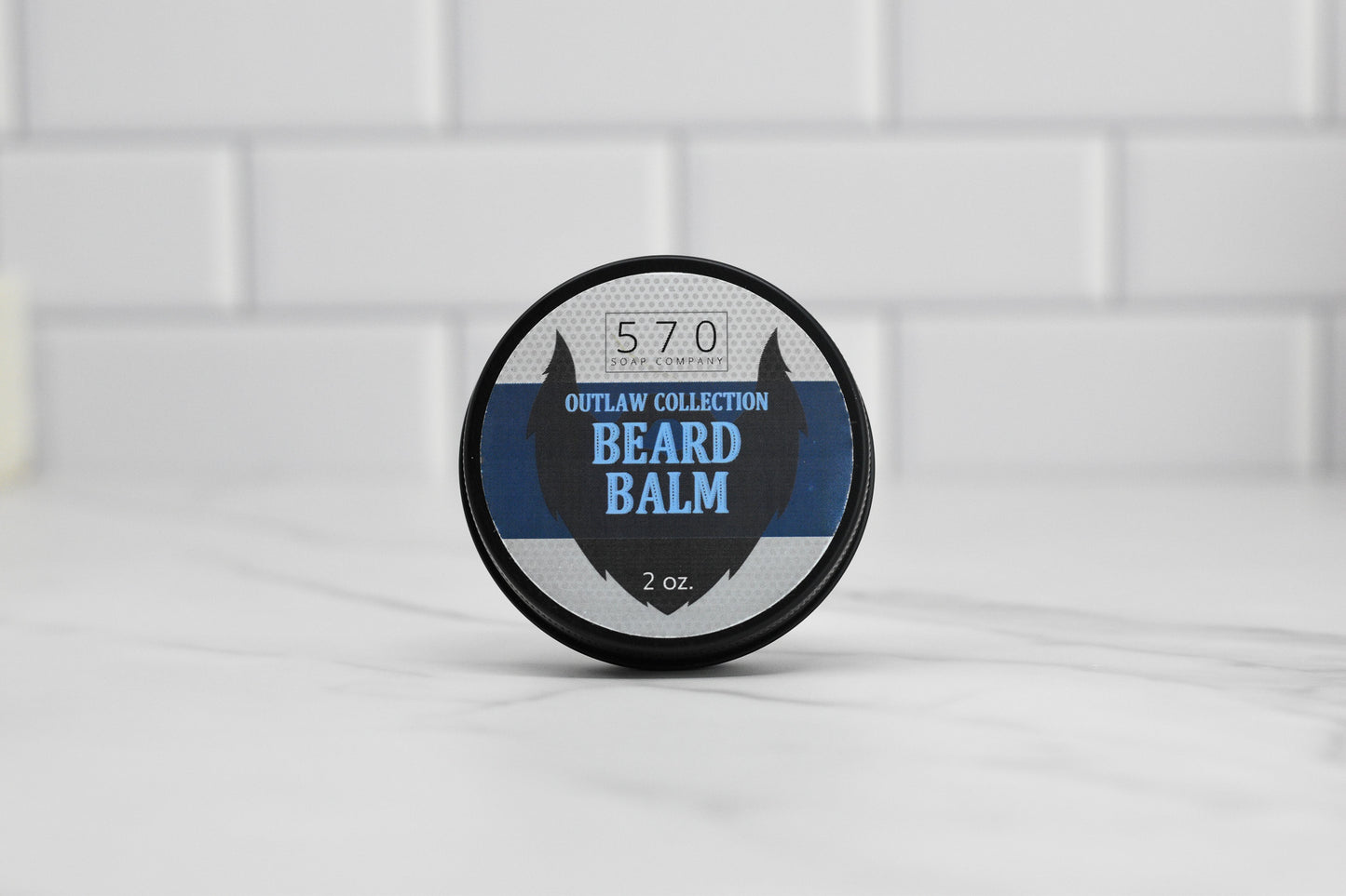 A round container of 570 Soap Co.'s Beard Balm - Choose your scent, featuring a natural touch for grooming beards and a hint of Kentucky Bourbon scent, rests on a marble surface against a backdrop of white tiles. The lid is adorned with a striking blue and black design accompanied by bold white text.