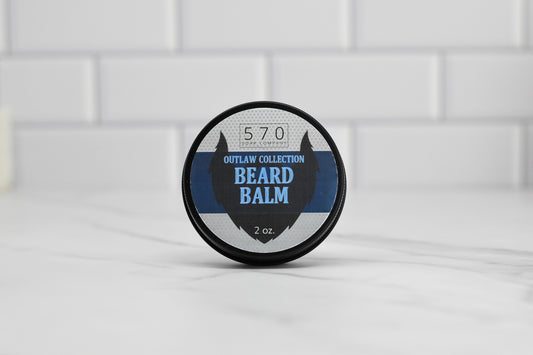 A round container of 570 Soap Co.'s Beard Balm - Choose your scent, featuring a natural touch for grooming beards and a hint of Kentucky Bourbon scent, rests on a marble surface against a backdrop of white tiles. The lid is adorned with a striking blue and black design accompanied by bold white text.