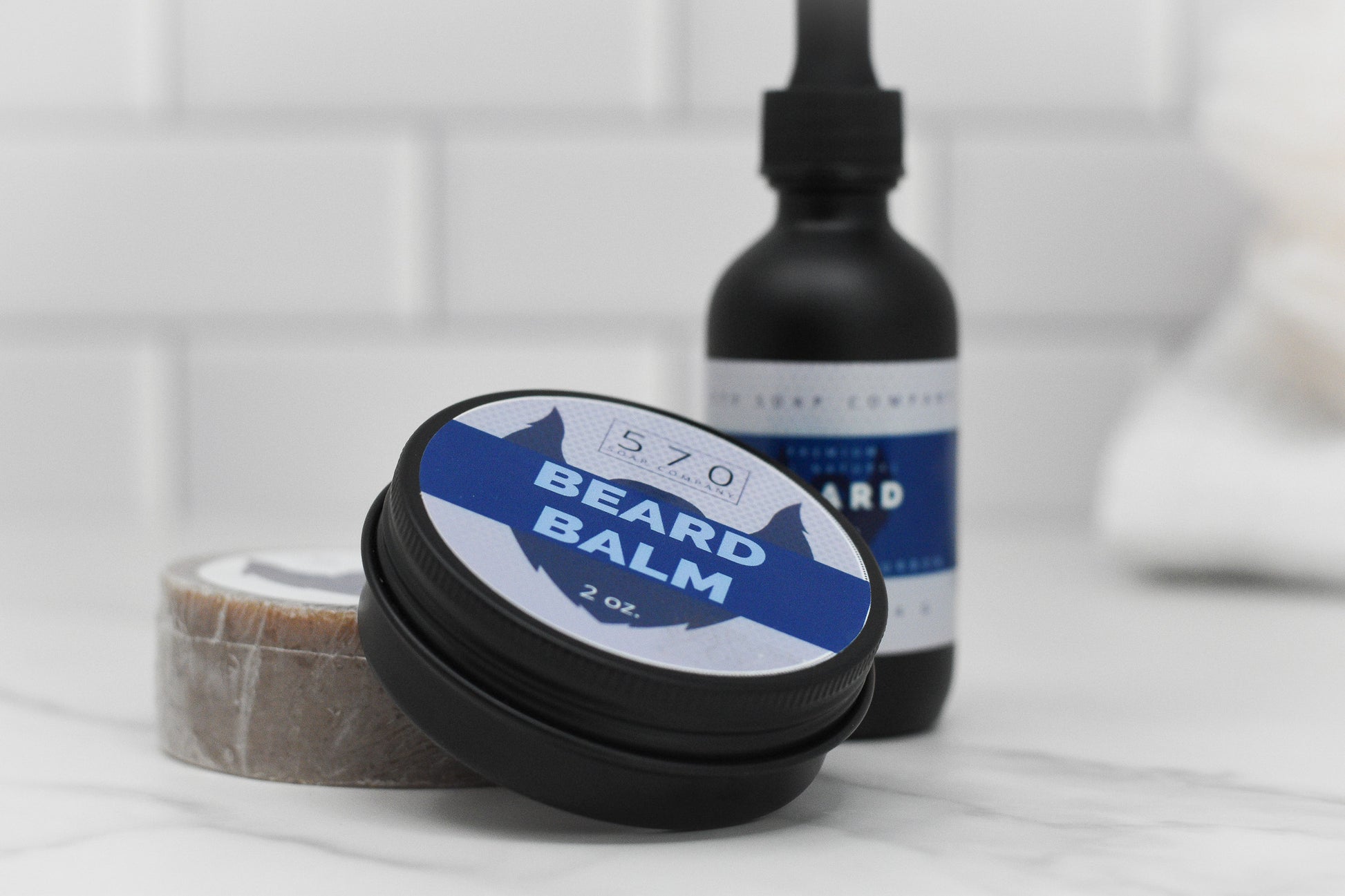 A 2 oz container of 570 Soap Co.'s Beard Balm, featuring the luxurious Kentucky Bourbon fragrance, is in the foreground, with a dropper bottle of beard oil positioned behind it. Placed on a white tiled surface, these grooming essentials offer an enhanced natural beard care experience.