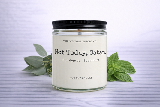 Candle jar labeled 'Not Today, Satan' with eucalyptus and spearmint leaves on a light background