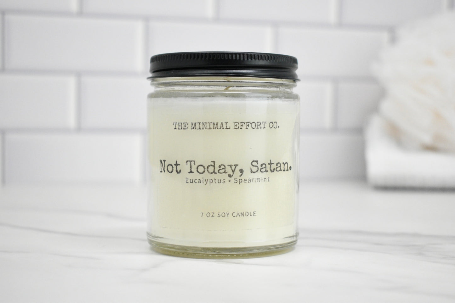 Candle jar labeled 'Not Today, Satan' by The Minimal Effort Co. on a white surface with a tiled wall background.
