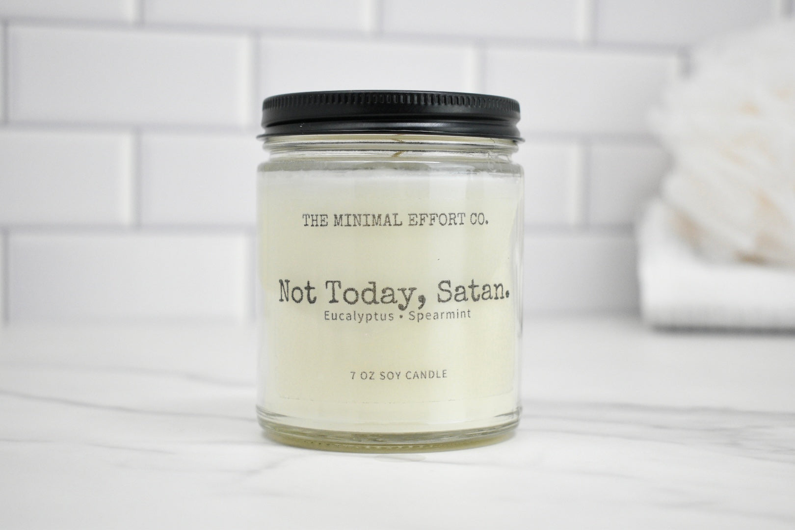 Candle jar labeled 'Not Today, Satan' by The Minimal Effort Co. on a white surface with a tiled wall background.