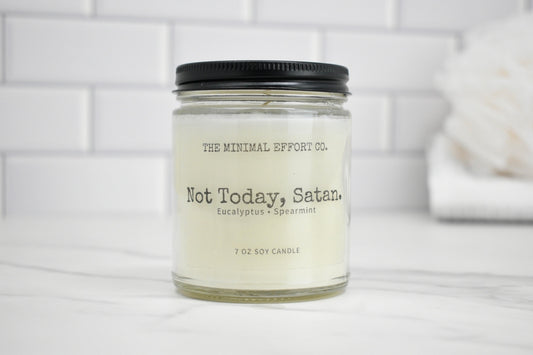 Candle jar labeled 'Not Today, Satan' by The Minimal Effort Co. on a white surface with a tiled wall background.