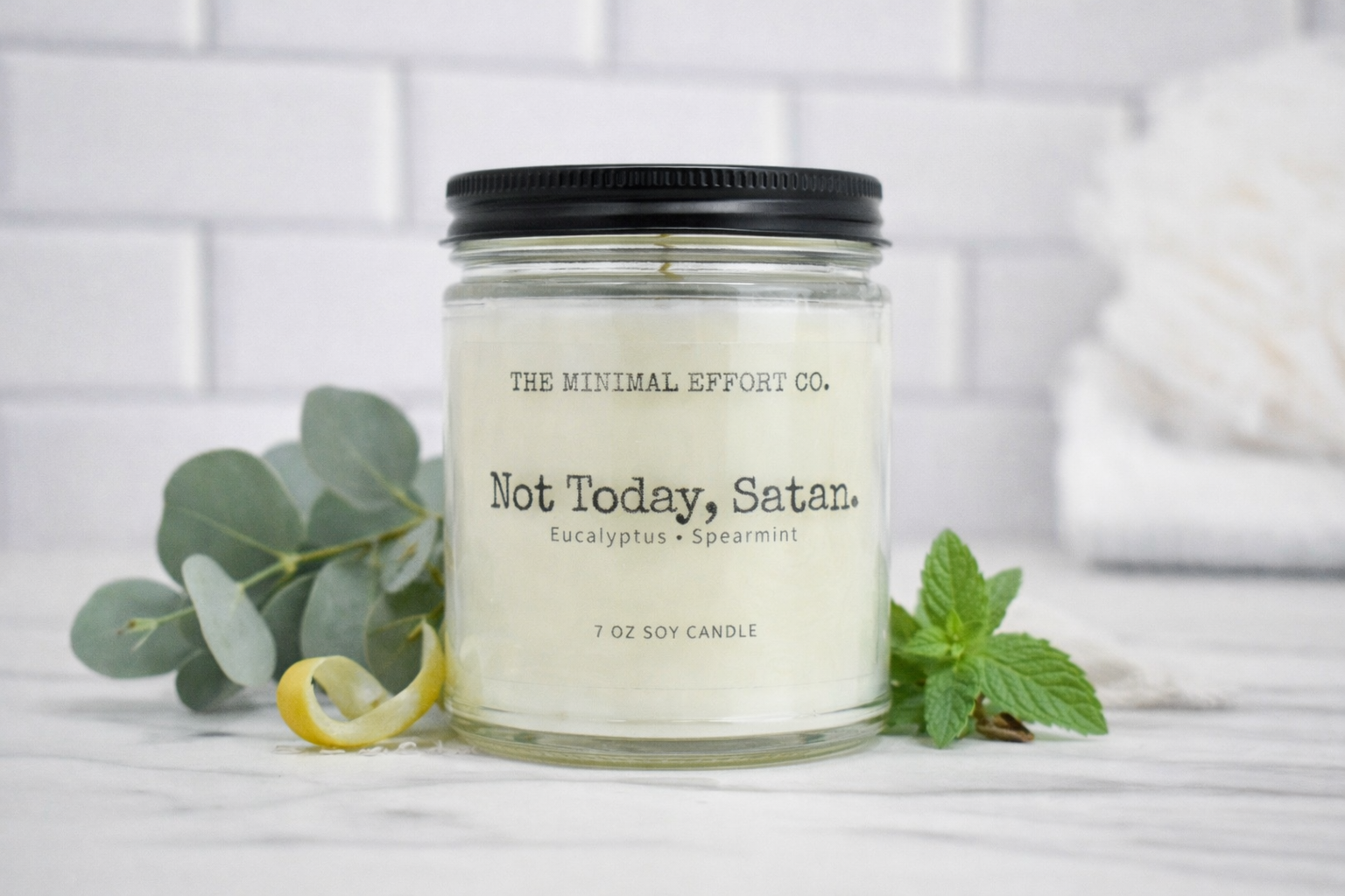 Candle jar labeled 'Not Today, Satan' from The Minimal Effort Co. on a white surface with greenery.