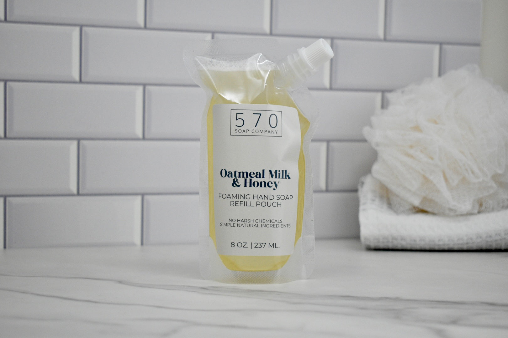 Oatmeal Milk & Honey 570 Soap refill pouch on a bathroom counter with a white tiled wall background.