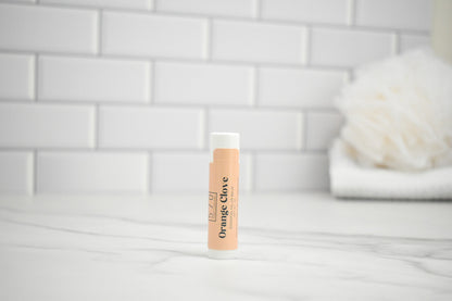 Lip balm with 'Orange Clove' flavor on a white surface with a tiled wall background