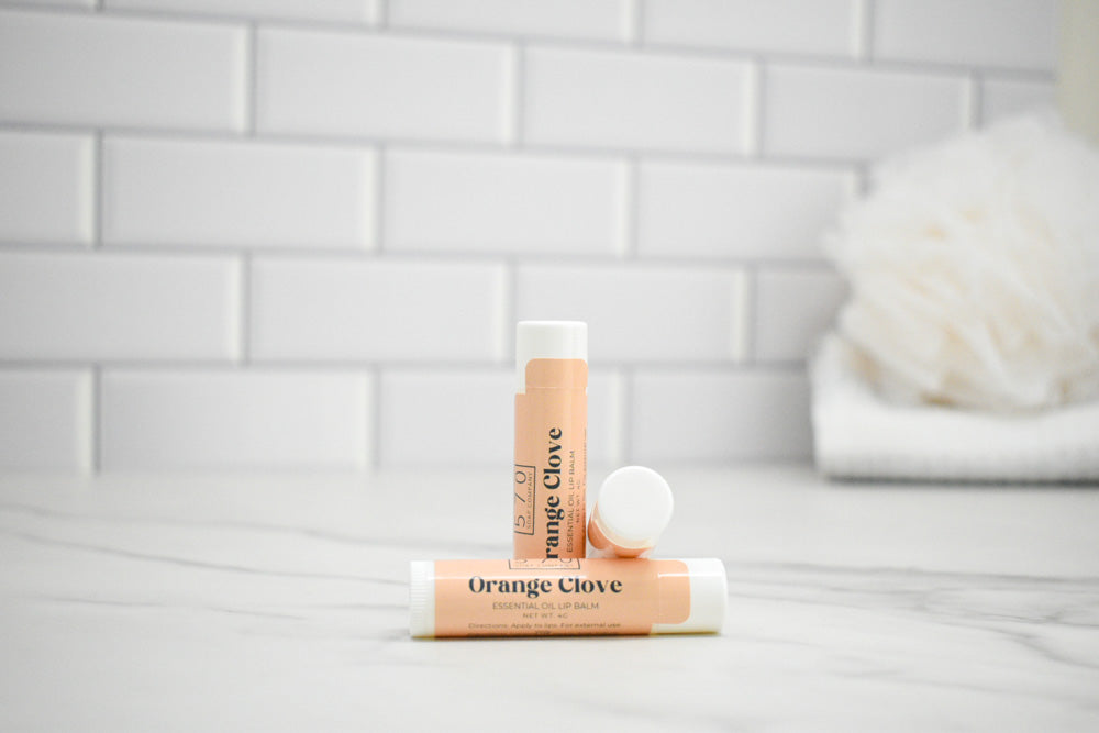 Two lip balm tubes labeled 'Orange Clove' on a white surface with a tiled wall background.