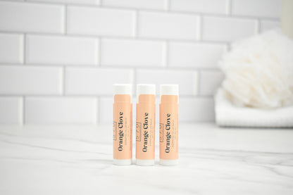 Three lip balm tubes labeled 'Orange Clove' on a white surface with a tiled wall background.