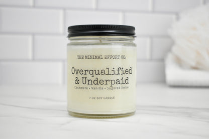 Candle jar labeled 'Overqualified & Underpaid' by The Minimal Effort Co. on a white surface with a tiled wall background.