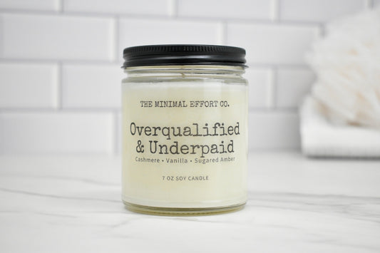 Candle jar labeled 'Overqualified & Underpaid' by The Minimal Effort Co. on a white surface with a tiled wall background.