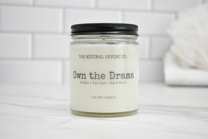 Candle jar labeled 'Own the Drama' by The Minimal Effort Co. on a white surface with a tiled wall background.