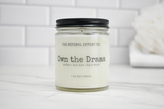 Candle jar labeled 'Own the Drama' by The Minimal Effort Co. on a white surface with a tiled wall background.