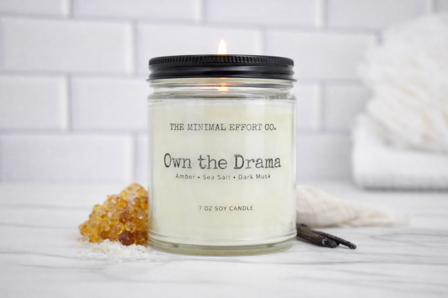 Candle in a glass jar labeled 'The Minimal Effort Co. Own the Drama' on a white surface with a tiled wall background.