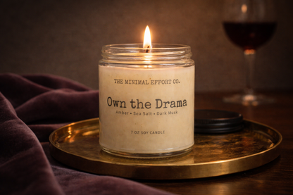 Candle labeled 'The Minimal Effort Co. Own the Drama' on a gold tray with a glass of red wine in the background.