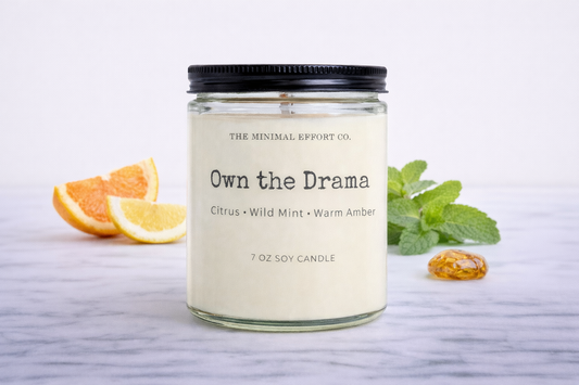 Candle jar labeled 'Own the Drama' with citrus and mint elements on a light background