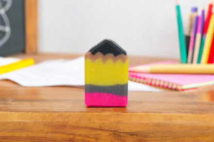 A multicolored pencil-shaped bar of soap from 570 Soap Co. is placed on a wooden desk, resembling a cheerful back-to-school surprise. The soap features layers of yellow, black, and pink. In the background, papers and colorful pencils rest in a container.