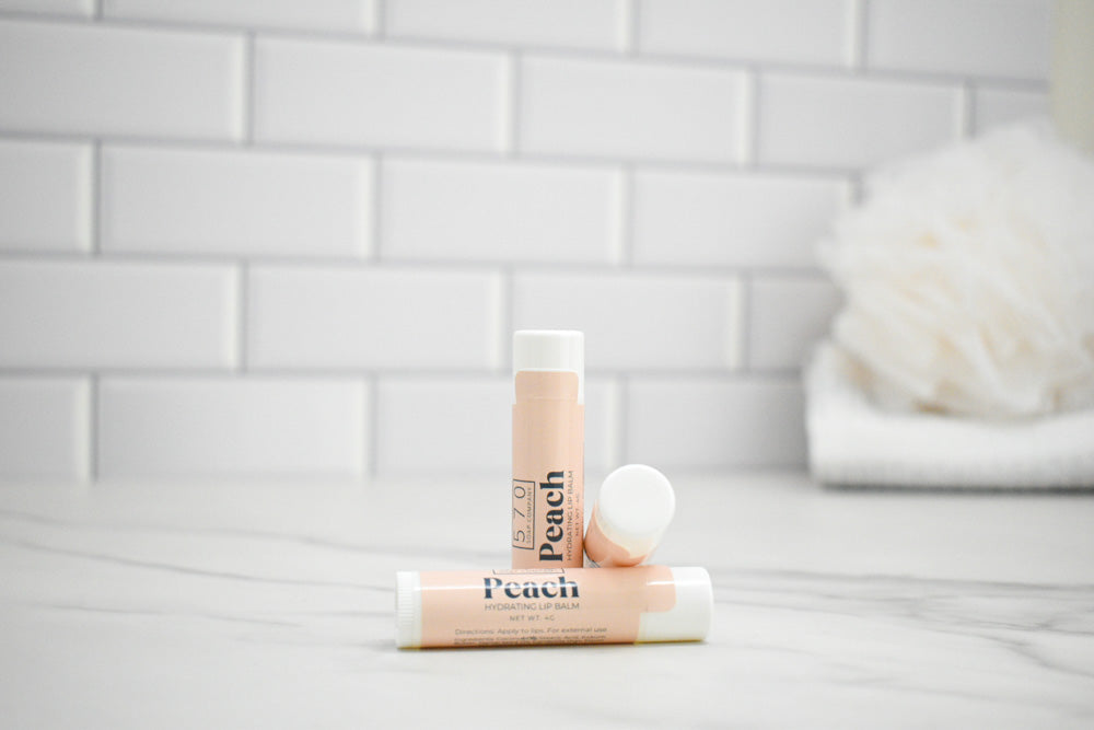 Two lip balms labeled 'Peach' on a bathroom counter with a tiled wall background.