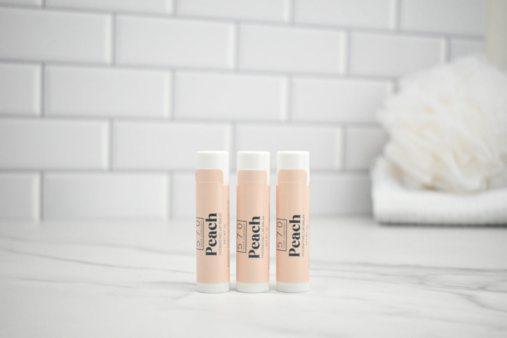 Three lip balm tubes labeled 'Peach' on a bathroom counter with a tiled wall background.