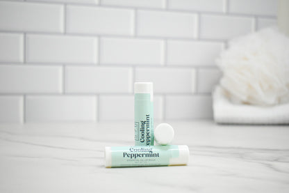 Two lip balm tubes labeled 'Cooling Peppermint' on a bathroom counter with a tiled wall background.