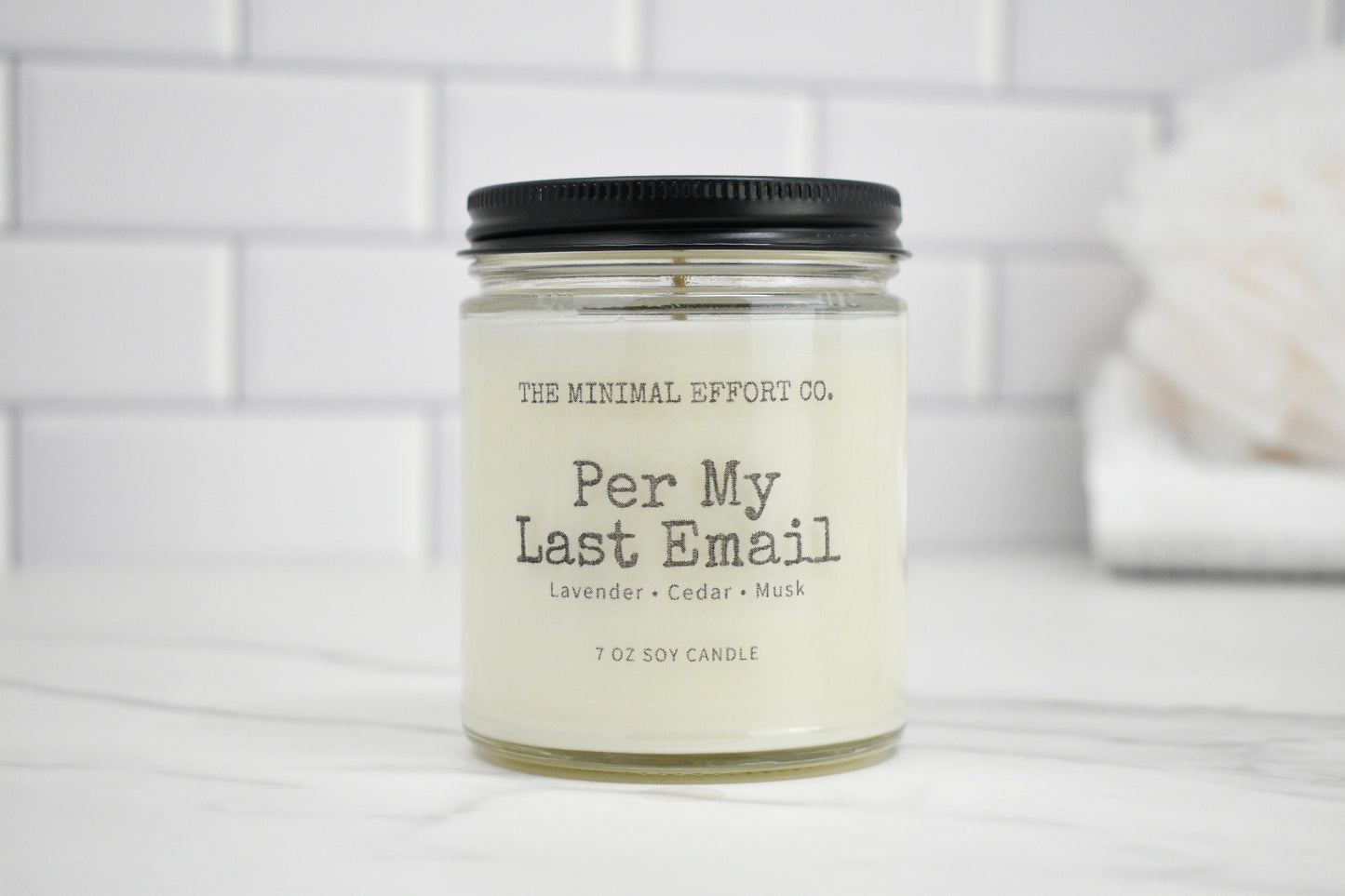 Candle jar labeled 'Per My Last Email' by The Minimal Effort Co. on a white surface with a tiled wall background.