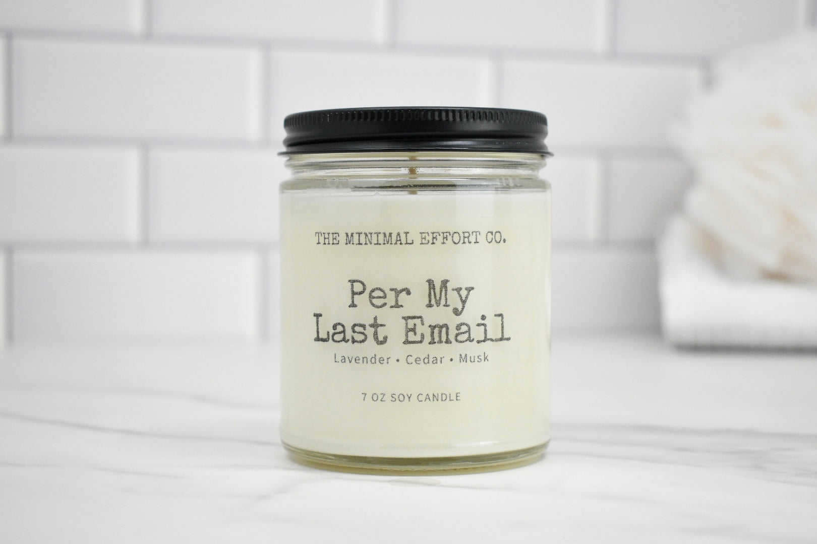 Candle jar labeled 'Per My Last Email' by The Minimal Effort Co. on a white surface with a tiled wall background.