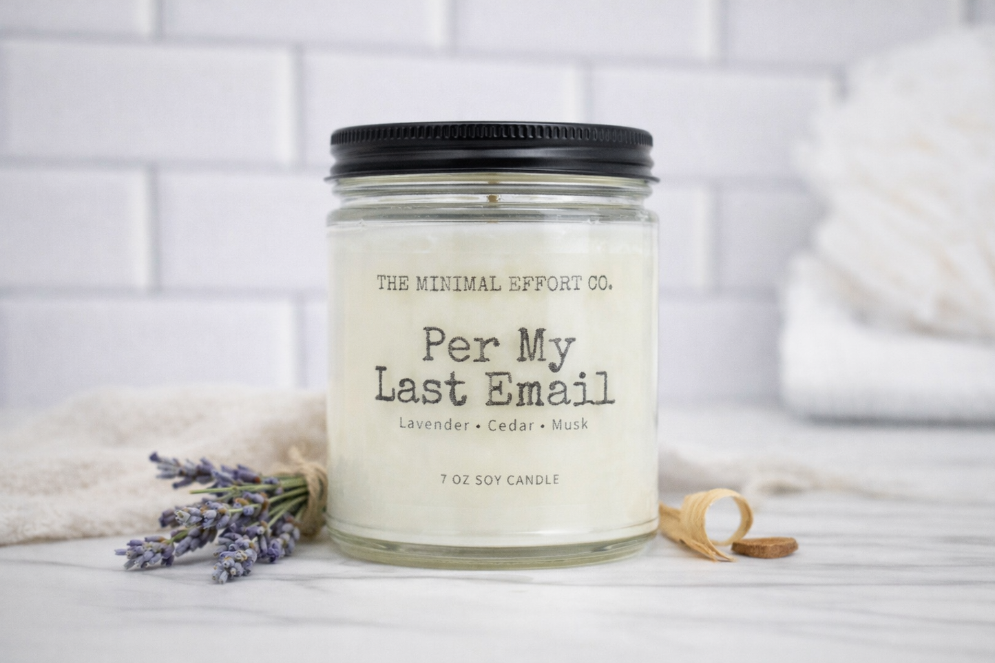 Candle labeled 'Per My Last Email' from The Minimal Effort Co. on a white surface with lavender and a wooden spoon.