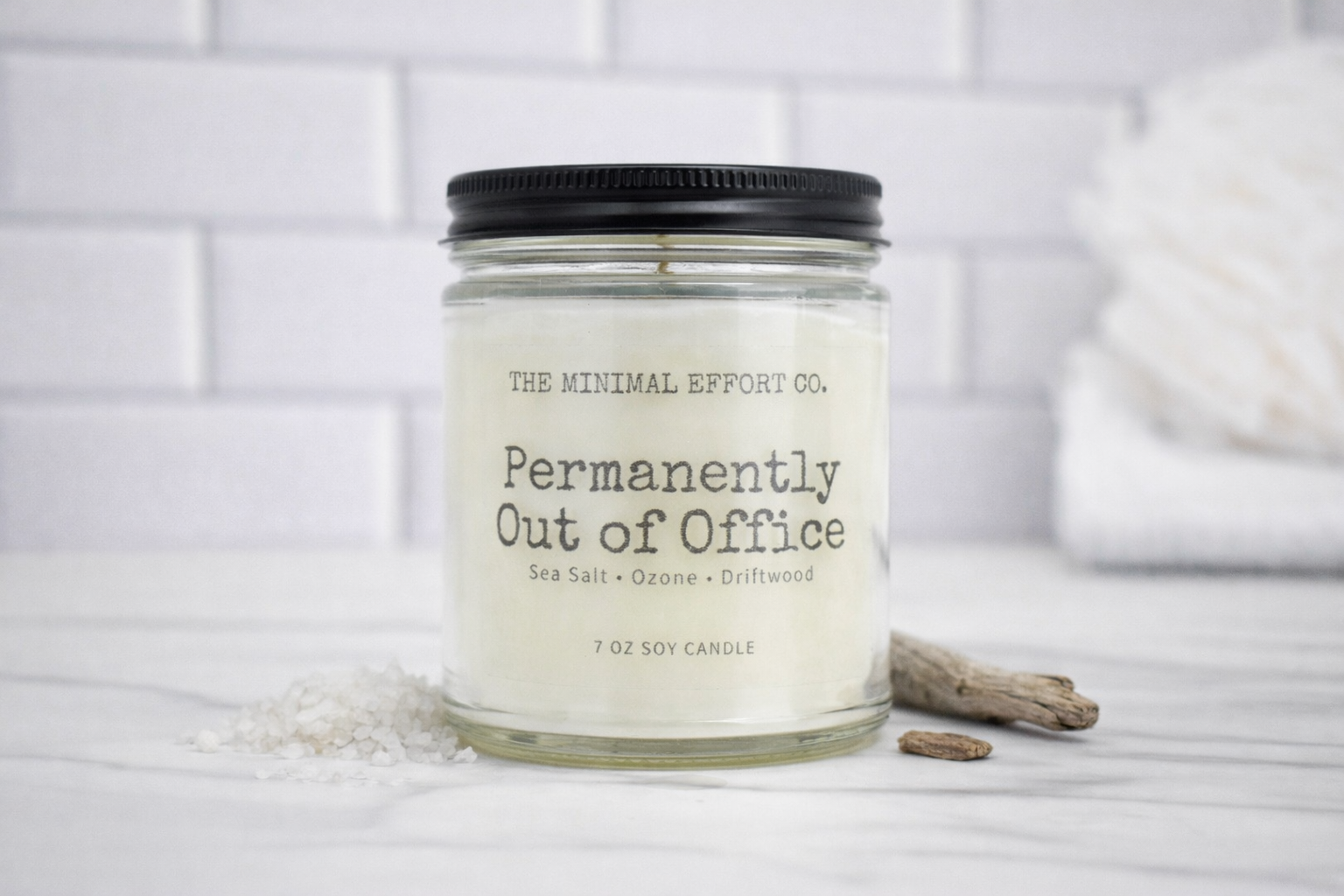 Candle labeled 'Permanently Out of Office' by The Minimal Effort Co. on a white surface with a neutral background.