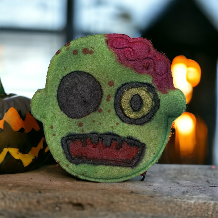 A close-up of a bath bomb from 570 Soap Co.'s Spooky Season collection, designed with a single eye, part of its surface revealing red color resembling a brain, and jagged patterns embodies the essence of Halloween. It rests on a wooden surface with a blurred pumpkin decoration in the background.