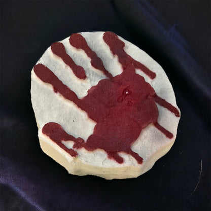 Against a dark fabric backdrop, a circular clay disk displays a textured red handprint, evoking the Spooky Season Bath Bombs by 570 Soap Co., with paint artfully dripping down the sides.