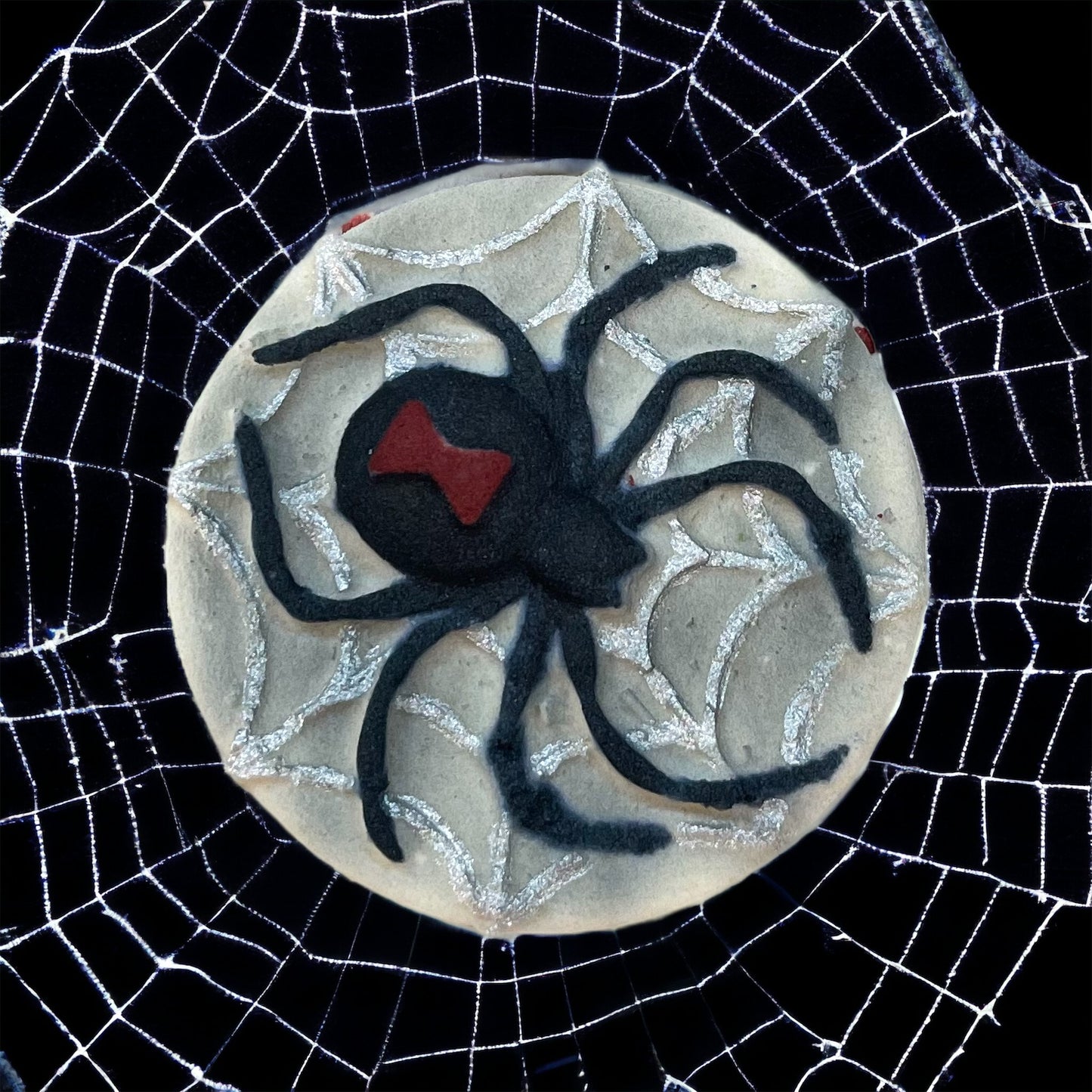 A round bath bomb adorned with a black spider design and a red marking on its back, surrounded by a web pattern. Perfect for Spooky Season, this bath bomb sits on an intricate white spiderweb against a black background, capturing the eerie charm of haunted nights. Presented by 570 Soap Co., it adds a mysterious touch to your bath experience.