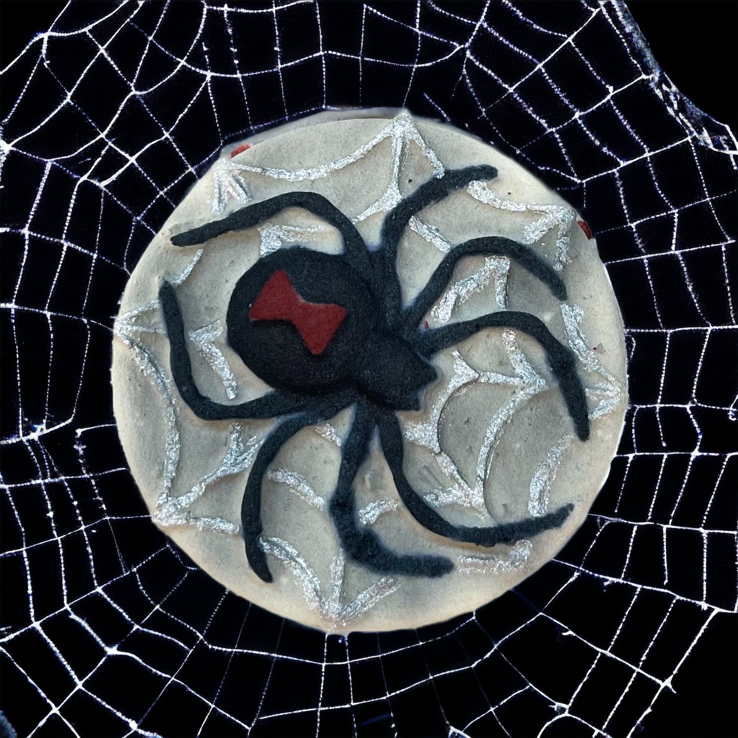 A round bath bomb adorned with a black spider design and a red marking on its back, surrounded by a web pattern. Perfect for Spooky Season, this bath bomb sits on an intricate white spiderweb against a black background, capturing the eerie charm of haunted nights. Presented by 570 Soap Co., it adds a mysterious touch to your bath experience.