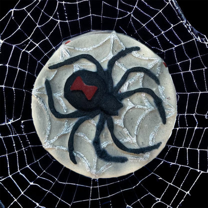 A round bath bomb adorned with a black spider design and a red marking on its back, surrounded by a web pattern. Perfect for Spooky Season, this bath bomb sits on an intricate white spiderweb against a black background, capturing the eerie charm of haunted nights. Presented by 570 Soap Co., it adds a mysterious touch to your bath experience.