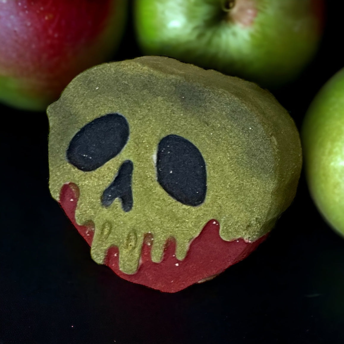 A red and green apple-shaped bath bomb with a painted design resembling a melting skull, part of the Spooky Season Bath Bombs collection by 570 Soap Co., sits against a dark background.