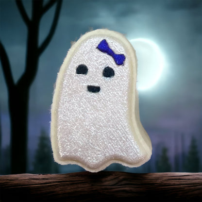 A Spooky Season Bath Bomb from 570 Soap Co., shaped like a charming ghost with a shimmering white surface and adorned with a tiny purple bow, sits on a wooden log. Capturing the essence of the season, the background showcases an eerie forest silhouetted against a full moon.