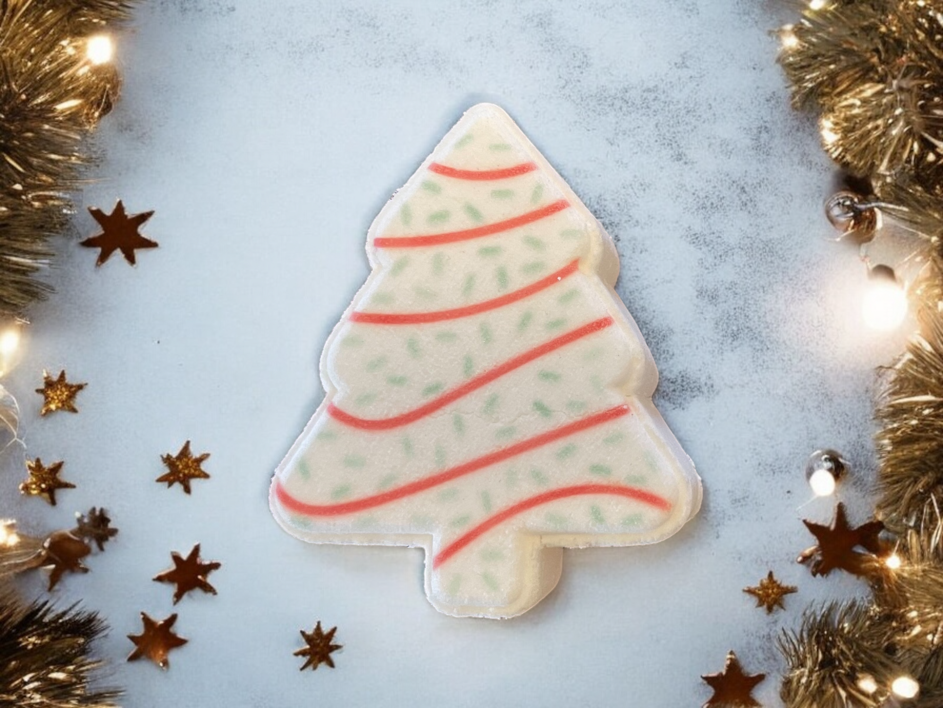 A Christmas tree-shaped cookie adorned with white icing, red swirl lines, and green dots sits amid gold tinsel and small star decorations against a blue backdrop. Twinkling lights enhance the festive atmosphere, similar to the sparkle found in Christmas Tree Cake Bath Bombs from 570 Soap Co.