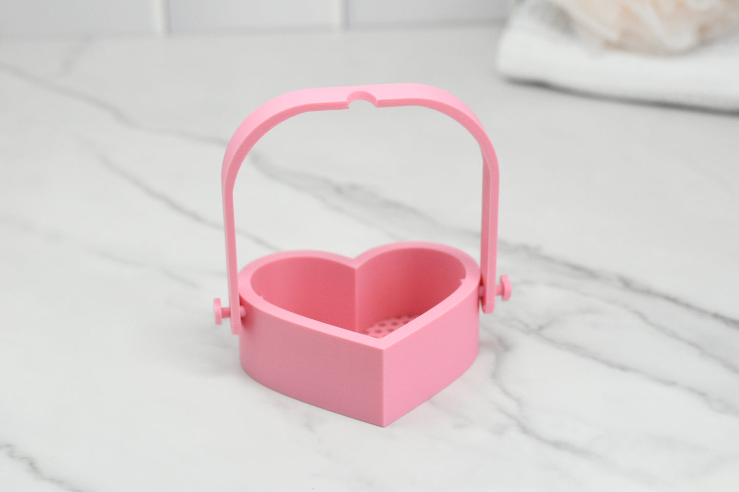 A pink, heart-shaped Bubble Bath Strainer with a handle from 570 Soap Co. sits on the marble countertop, evoking the charm of a luxurious oasis in your kitchen.