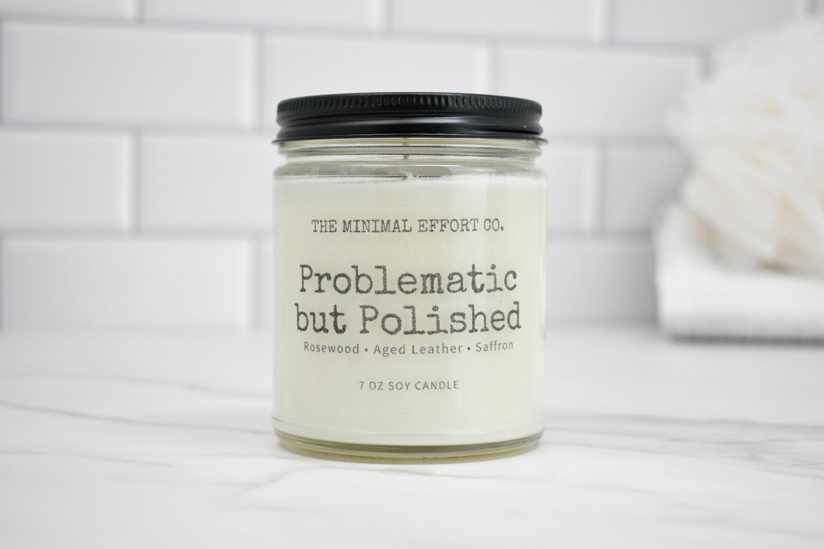 Candle jar labeled 'Problematic but Polished' by The Minimal Effort Co. on a white surface with a tiled wall background.