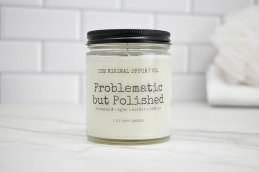 Candle jar labeled 'Problematic but Polished' by The Minimal Effort Co. on a white surface with a tiled wall background.