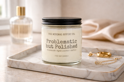 Candle labeled 'Problematic but Polished' by The Minimal Effort Co. on a marble tray with a bottle and jewelry in the background.