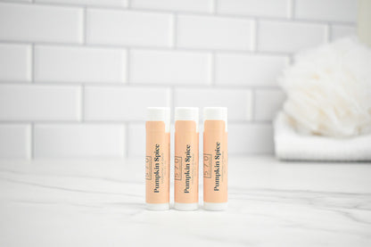 Three lip balm tubes labeled 'Pumpkin Spice' on a white surface with a tiled wall background.