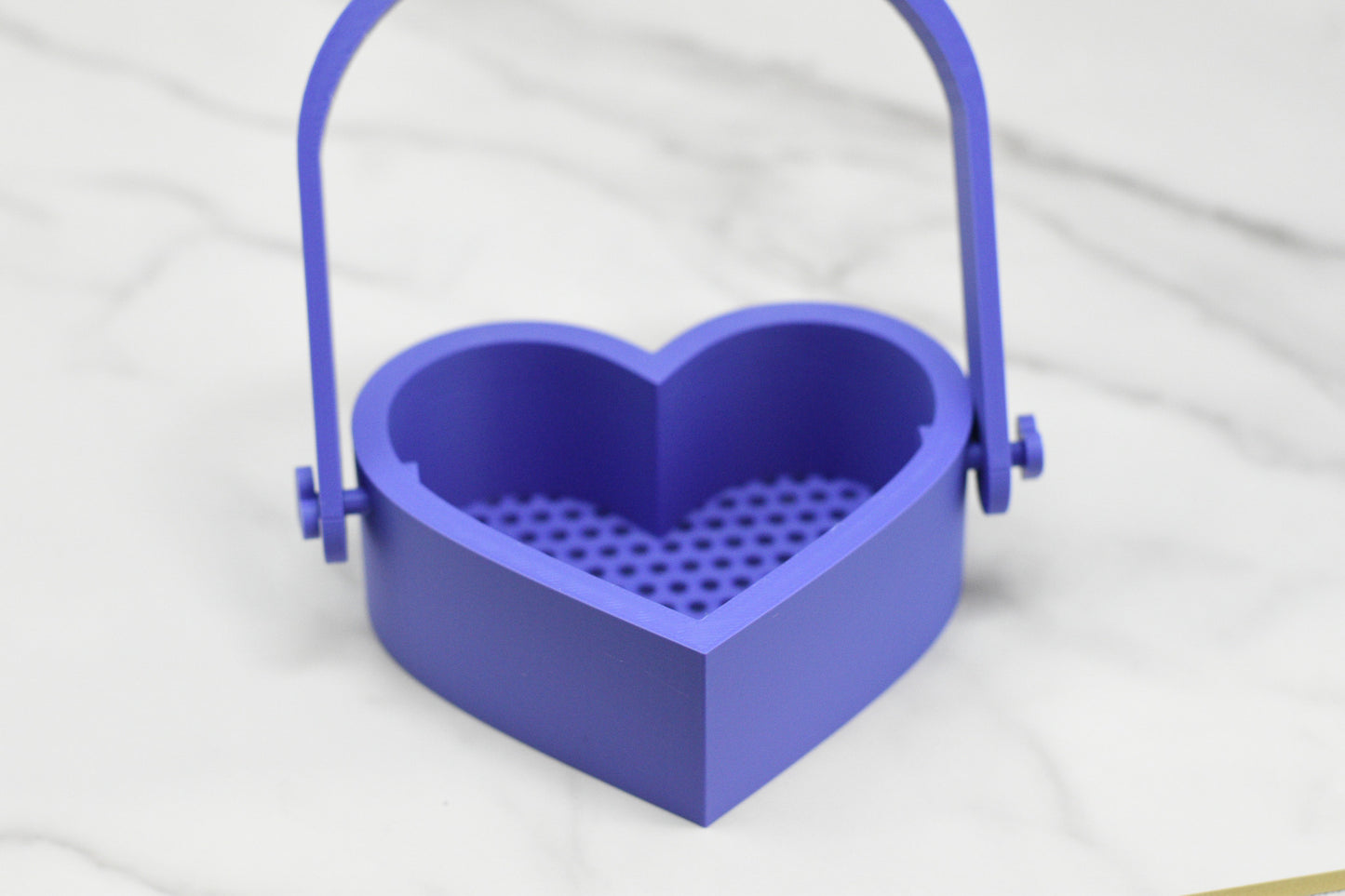 The Bubble Bath Strainer by 570 Soap Co. is a purple, heart-shaped container with a handle and a perforated bottom, crafted to enhance your Foamy Bubble Experience. Resting on a marble surface, it elevates any setting into a luxurious haven of relaxation and style.