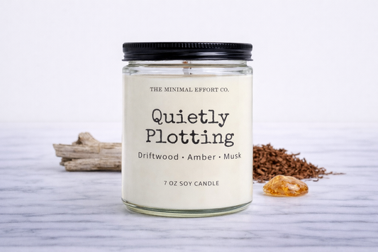 Candle jar labeled 'Quietly Plotting' with natural elements on a light background
