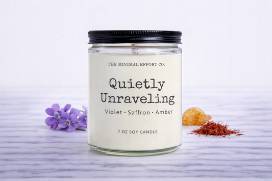 Candle labeled 'Quietly Unraveling' with ingredients displayed on a white background