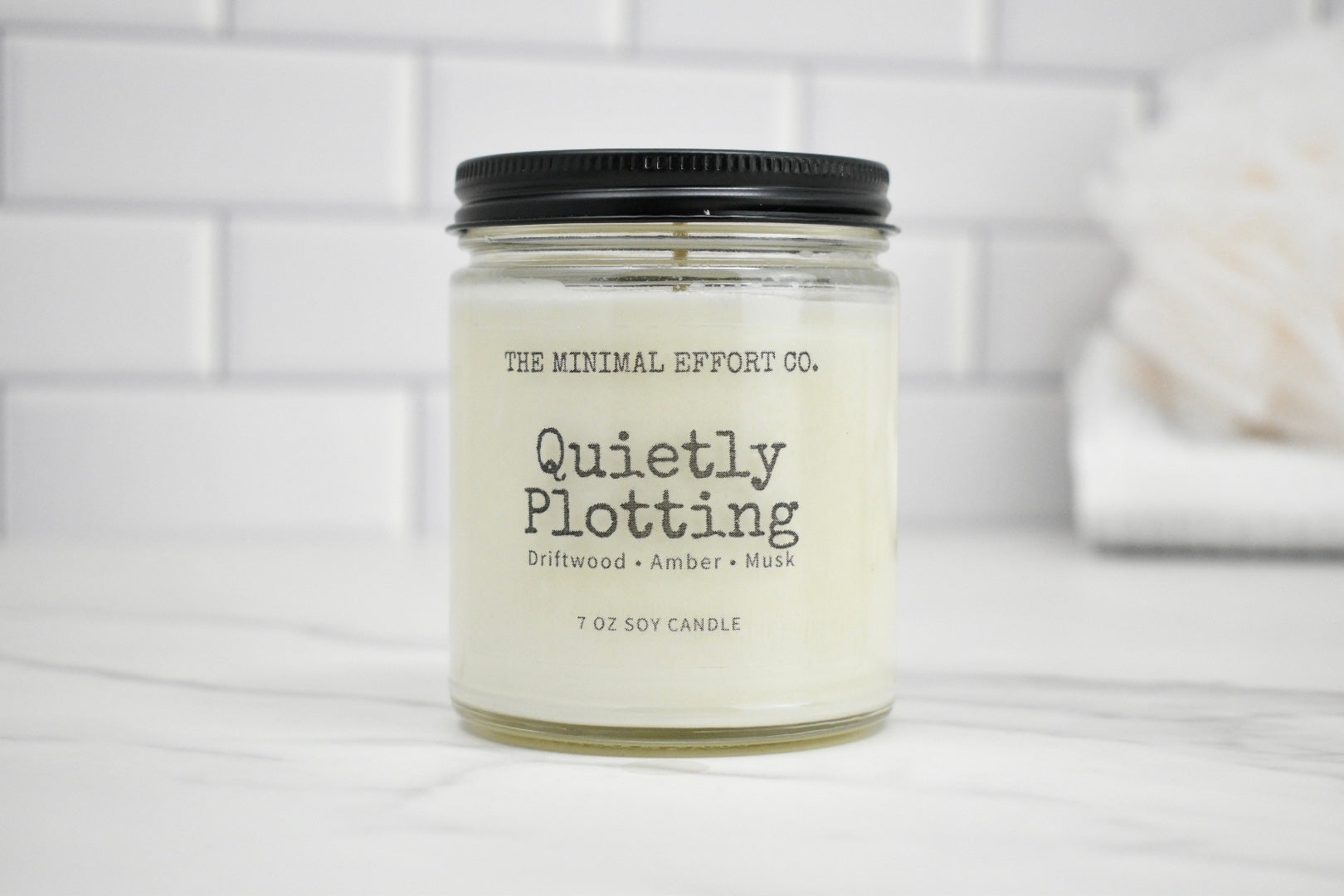 Candle jar labeled 'Quietly Plotting' by The Minimal Effort Co. on a white background