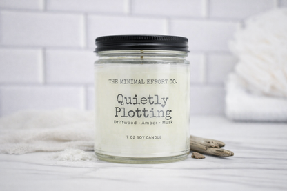 Candle with 'The Minimal Effort Co.' branding on a white surface with a blurred background