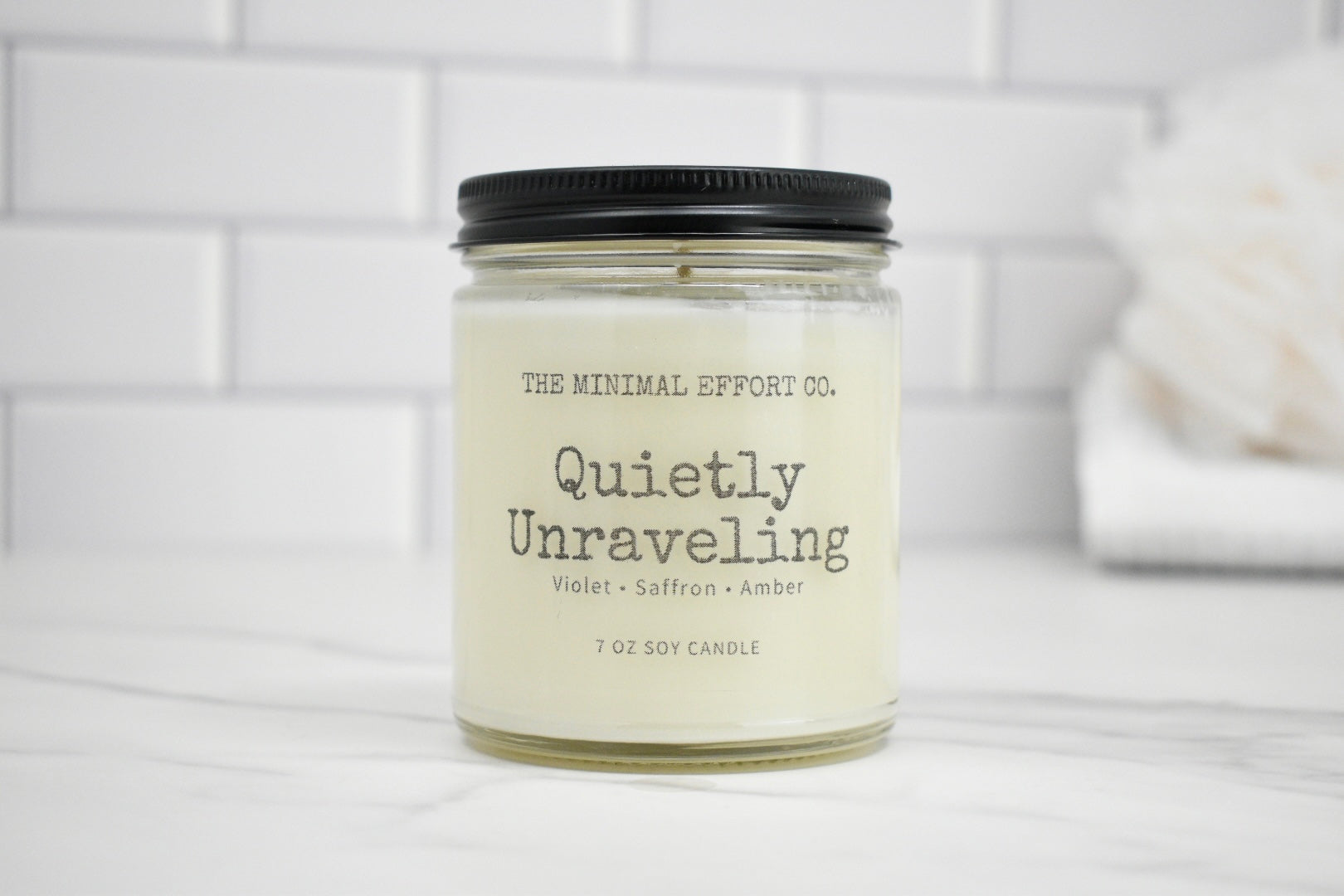 Candle jar labeled 'Quietly Unraveling' by The Minimal Effort Co. on a white surface with a tiled wall background.